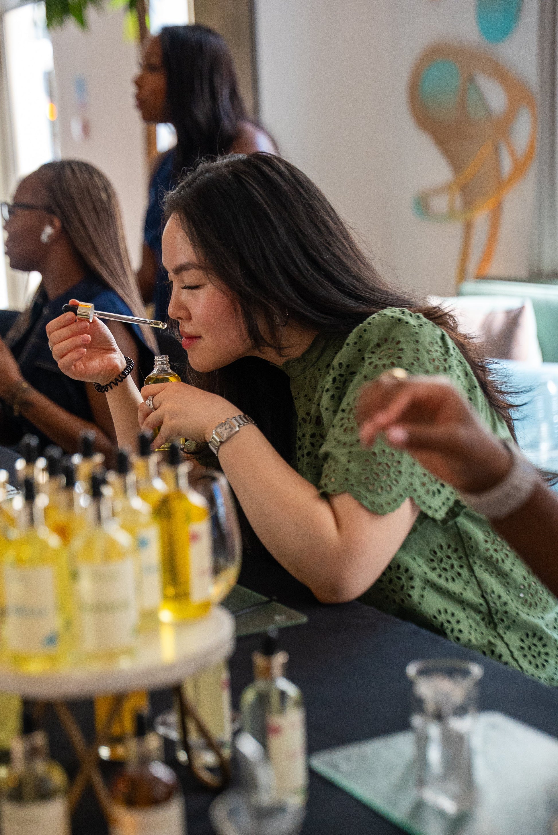 A woman in a green patterned top samples a fragrance from a small glass bottle at a table filled with bottles and glassware, in a bright indoor setting at Reverie.